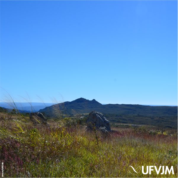 Fotografia mostra uma paisagem com céu azul sem nuvens, montanhas ao fundo e, à frente, uma vegetação rasteira com pedras. No canto inferior esquerdo está a assinatura da Dicom e, no direito, a logomarca da UFVJM.