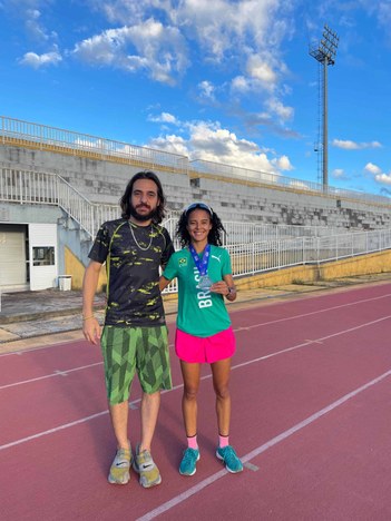 Livya se encontra em pé ao lado do Professor Danilo segurando sua medalha de prata em uma pista de atletismo. Ela veste uma roupa com cores verdes e azul, enquanto Danilo veste cores verde e preto.